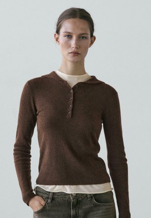 Giovane donna con pelle chiara e capelli raccolti indossa un maglione marrone a maniche lunghe sopra una camicia beige, posando con una mano nella tasca dei jeans.