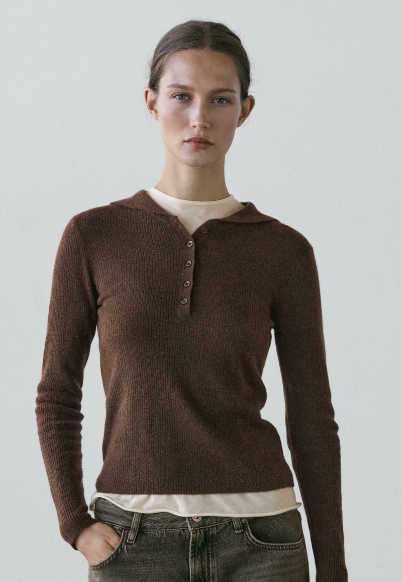 Young woman with light skin and pulled-back hair wears a brown long-sleeve knit top over a beige shirt, posing with one hand in jean pocket.