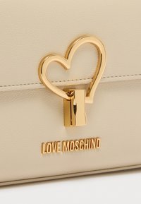 Μπεζ δερμάτινη τσάντα χειρός με χρυσό κούμπωμα σε σχήμα καρδιάς και το "LOVE MOSCHINO" σε χρυσά γράμματα. Λεία υφή με ελάχιστες ραφές.