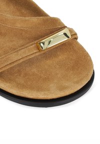 Tan suede sandal with a gold metallic accent. The design features crossed straps and a textured surface, set on a black sole.