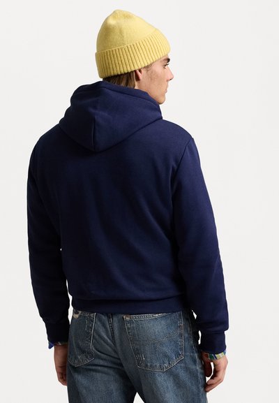 Sudadera con capucha azul marino de corte relajado, hecha de una tela suave. Combinada con jeans azul claro y un gorro de canalé amarillo.