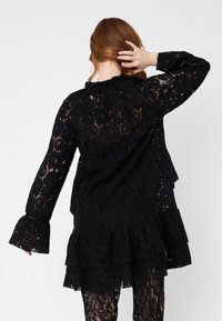 Rochie neagră din dantelă, cu mâneci lungi tip clopot, decolteu volănaș și poale în straturi. Model floral complex pe întreaga țesătură.