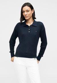 Σκούρο μπλε πλεκτό πόλο πουλόβερ με υφή, που διαθέτει γιακά, τρία χρυσά κουμπιά και ριμπ μανσέτες και τελείωμα. Φορεμένο με λευκό παντελόνι.