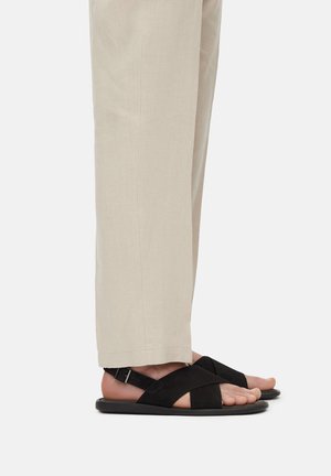 Czarne sandały zamszowe z krzyżującym się paskiem i regulowaną klamrą na wąskiej, płaskiej podeszwie, noszone z jasnymi, prostymi spodniami.