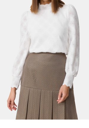 Vrouw draagt een witte, gestructureerde blouse met lange mouwen met pofschouders en een hooggetailleerde bruine geruite plissérok.