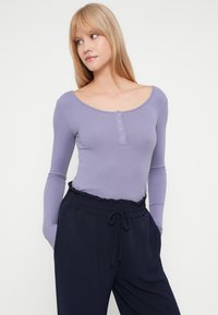 Νεαρή γυναίκα με ίσια ξανθά μαλλιά, φορεμένη με ένα λειαντό μωβ μπλουζάκι με μακριά μανίκια και ναυτικές παντελόνες με κορδόνια, με τα χέρια στις τσέπες, κοιτάζοντας αριστερά.