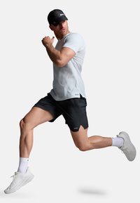 Camiseta deportiva gris claro, pantalones cortos negros y zapatillas de correr blancas. La figura está en una postura dinámica de carrera, llevando una gorra negra.