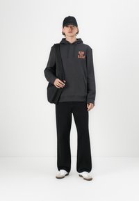 Sudadera gris con "RIDE THE RAILS" en naranja, pantalones de pierna ancha negros y zapatillas blancas. Una gorra negra y una bolsa completan el atuendo.