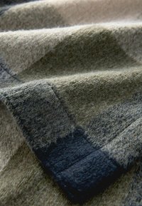 Woolen textile with a soft, textured surface. Features earthy green, beige, and dark gray tones. Includes a grid-like pattern and folded edges.