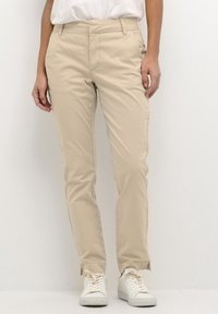 Beige slim-fit trousers with a smooth texture, featuring front pockets and zipper details at the ankles, paired with white sneakers.
