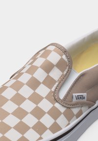 Αθλητικό παπούτσι τύπου slip-on με μοτίβο καρό σε μπεζ και άσπρο, ελαστικές πλευρικές λεπτομέρειες και ετικέτα με το λογότυπο Vans στο πλαϊνό πάνελ.