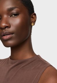 Złote geometryczne kolczyki-stud w uszach modelki o ciemnej karnacji, która ma na sobie brązową bluzkę bez rękawów, charakteryzującą się gładką fakturą i prostym wzornictwem.