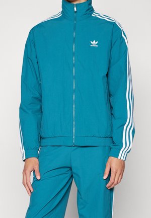 Türkisfarbener Adidas-Trainingsanzug mit weißen drei Streifen an Ärmeln und Hosen, Jacke mit Frontreißverschluss und Logo auf der linken Brust.