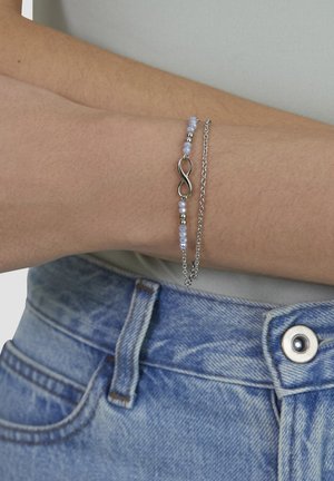 Wrist with layered silver bracelets, one with small light blue beads and an infinity symbol, wearing light blue denim jeans and a gray shirt.