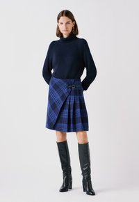 Maglione a collo alto lavorato a maglia di colore navy abbinato a una gonna avvolgente a quadri blu con fibbia nera, pieghe e stivali neri fino al ginocchio.