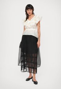 Valge volangiga topp, millel on sobiv vöökoht, koos pika musta läbipaistva seeliku ja lillemustritega. Mustad lapikesed kingad täiendavad riietust.