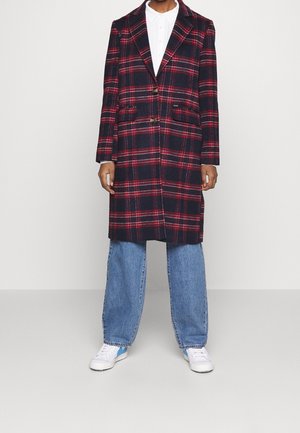 Personne portant un long manteau à carreaux bleu marine et rouge, une chemise blanche à col, un jean bleu ample et des baskets blanches, debout sur un fond clair.