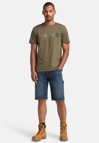 Camiseta de algodón verde oliva con el logo "Timberland", combinada con pantalones cortos de mezclilla azules y botas de trabajo marrones con cordones amarillos y una superficie texturizada.