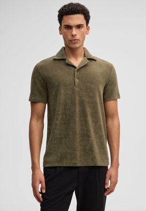 Mężczyzna o kręconych włosach, ubrany w oliwkowo-zieloną koszulkę polo z krótkim rękawem i czarne spodnie, stojący na jednolitym jasnym tle.