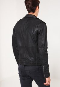 Man som bär en åtsittande svart läderjacka och mörkgrå jeans, vänd bort från kameran mot en enfärgad ljus bakgrund.