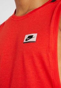 Nahaufnahme einer Person, die ein rotes ärmelloses Sportshirt mit einem kleinen schwarz-weißen Logo auf der Brust trägt.