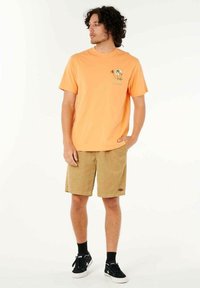 Helles orange Baumwoll-T-Shirt mit einem Palmbaum-Graphic; kombiniert mit beigen Cordshorts und schwarzen Sneakers mit weißen Sohlen und schwarzen Socken.