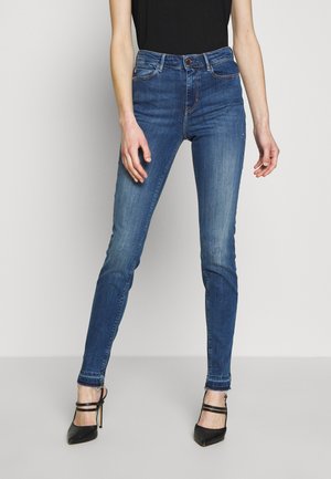 Frau trägt enge blaue Skinny-Jeans aus Denim und schwarze High-Heels, steht vor einem schlichten hellen Hintergrund.