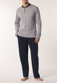 Uomo in camicia henley a maniche lunghe grigia chiara e pantaloni larghi blu scuro, a piedi nudi, su uno sfondo neutro.