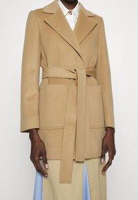 Frau trägt einen wollenen Mantel in Camel mit Gürtel und aufgesetzten Taschen über einem weißen Hemd und einem beigen Rock mit blauen Streifen.