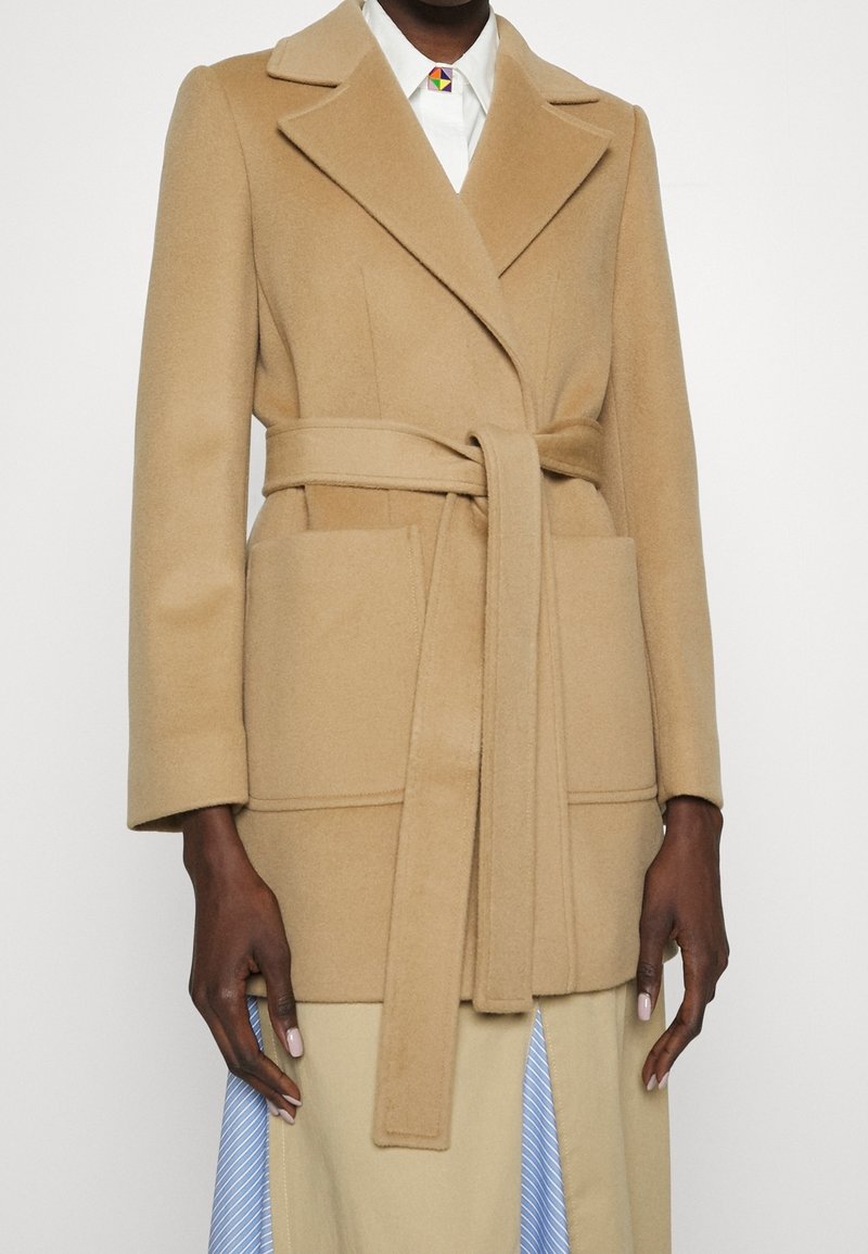 Frau trägt einen wollenen Mantel in Camel mit Gürtel und aufgesetzten Taschen über einem weißen Hemd und einem beigen Rock mit blauen Streifen.