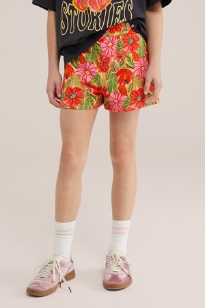 Person iført lyserøde og røde blomstrede shorts, hvide sokker, lyserøde sneakers og en sort t-shirt med grafik og delvist synlig tekst.