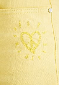 Una tela amarilla con un acabado texturizado presenta un corazón y un símbolo de paz bordados en un amarillo ligeramente más oscuro. Hay acentos cosidos alrededor del diseño.