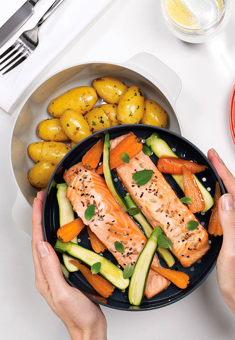 Zwarte schaal met twee zalmfilets, oranje wortelen en groene courgettes, gegarneerd met kruiden. Aan de zijkant een witte kom met gele baby-aardappelen.