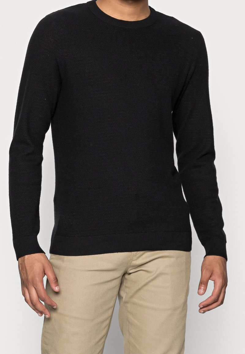 Pull noir à manches longues avec un col côtelé et un motif tricoté texturé, porté avec un pantalon beige clair.