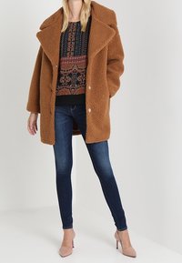 Cappotto teddy marrone con ampio bavero, tessuto testurizzato e vestibilità oversize; abbinato a jeans scuri e scarpe con tacco nudo.