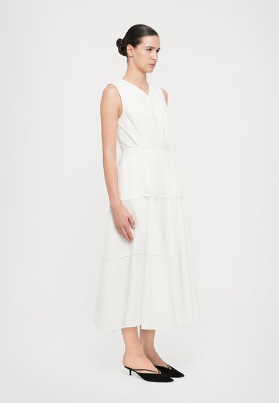 White, sleeveless dress with layered design, flowing skirt, v-neck, and soft texture. Worn with black, pointed-toe, heeled mules.