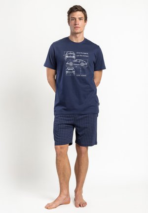 Joven de pie descalzo, con camiseta gráfica azul marino con dibujos de coches y pantalones cortos a cuadros azul marino sobre fondo blanco.