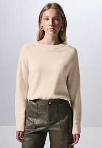 Pull en tricot beige avec un col rond et des poignets côtelés, associé à un pantalon en cuir vert foncé avec une fermeture à bouton et des poches latérales.