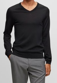 Černý dlouhý svetr s výstřihem do V vyrobený z hladkého úpletu, kombinovaný se světle šedými kalhotami. Svetr má přiléhavý střih a žebrované manžety.