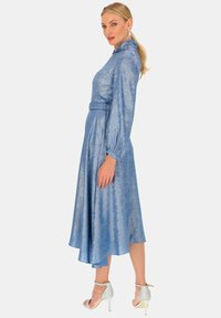 Niebieska satynowa sukienka z długim rękawem, wysokim kołnierzem, paskiem w talii i zwiewnym dołem. Noszona z srebrnymi szpilkami na kostkę. Gładka tekstura.