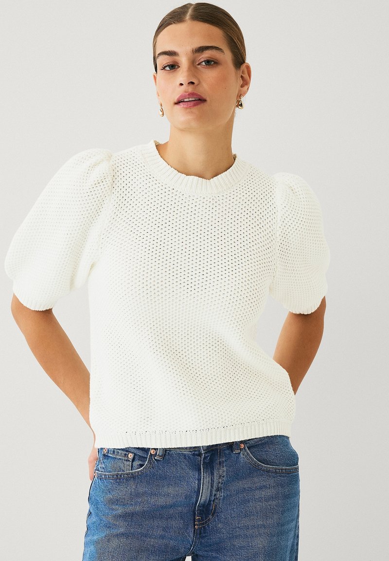 Maglione bianco a maglia con maniche corte a sbuffo, colletto e orlo a coste, indossato con jeans blu. Presenta un motivo a trama aperta testurizzata.