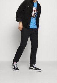 Chaqueta de pana negra, camiseta gráfica azul con diseño de palmera, pantalones negros y zapatillas altas negras con detalles en blanco.