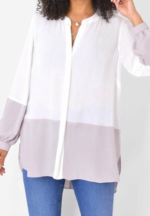 Frau trägt eine lockere, langärmlige Color-Block-Bluse in Weiß und Hellgrau mit blauer Jeans, ergänzt durch geschichtete silberne Halsketten.