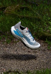 Chaussure de course Salomon avec un dessus gris et bleu, en maille texturée et logo bien visible ; semelle légère avec un design de crampons robuste.
