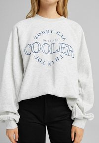 Grå sweatshirt med texten "Förlåt Bae men jag är MER KALL än du" i marinblått; har raglanärmar och resårvidda ärmslut; matchas med svarta byxor.