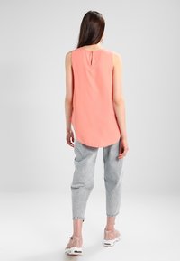 Žena s dlouhými hnědými vlasy, oblečená ve bezramenném korálovém topu, světle šedých džínách a růžových teniskách, stojící zády k šedému pozadí.