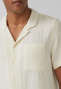 Lichtbeige shirt met korte mouwen en een ontspannen kraag, voorzak en gekreukelde structuur. Gemaakt van zachte, lichte stof.