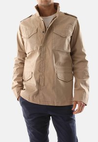 Giacca in cotone beige con colletto alto, due tasche sul petto e due tasche frontali. Presenta bottoni a pressione e una vestibilità leggermente rilassata.