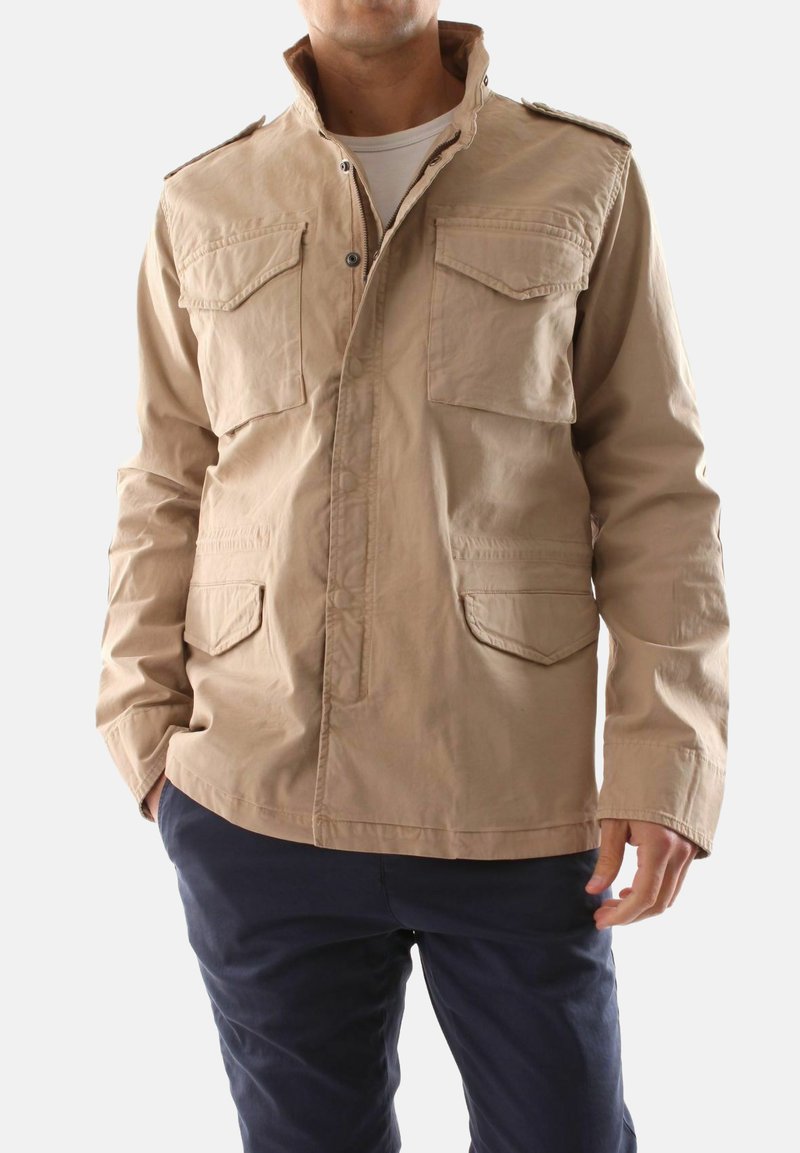 Giacca in cotone beige con colletto alto, due tasche sul petto e due tasche frontali. Presenta bottoni a pressione e una vestibilità leggermente rilassata.