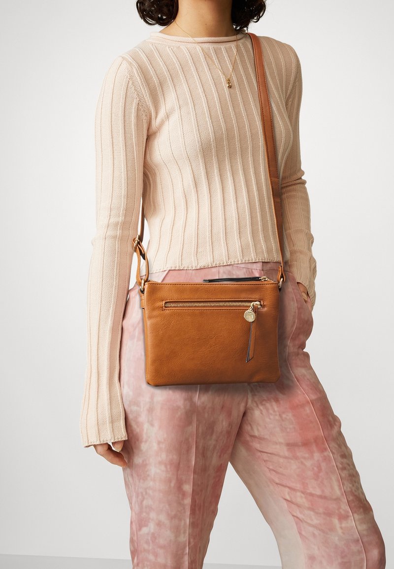 Kvinde iført en ribbet beige sweater og rosa tie-dye bukser med en tan læder crossbody taske med gulddetaljer på lynlåsen.